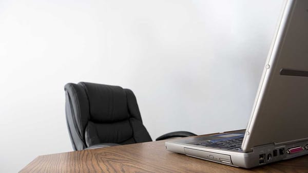 An empty office chair in a featureless room at a table in front of a laptop computer