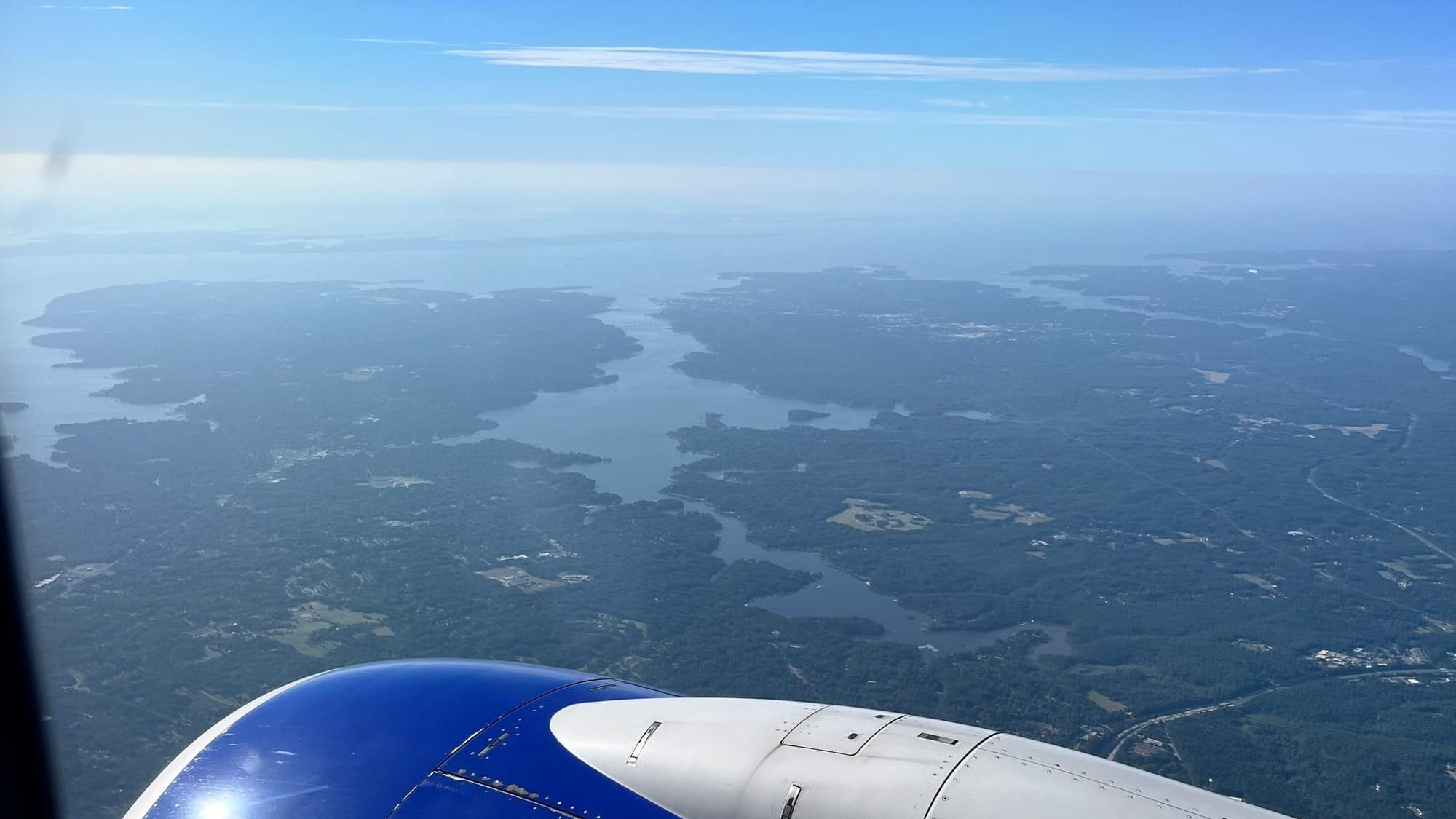 The top of a jet engine in the foreground, the left part of it painted blue, and beyond and below an inlet snaking through green land to a coastline full of other inlets, and the sea and haze and sky beyond. 