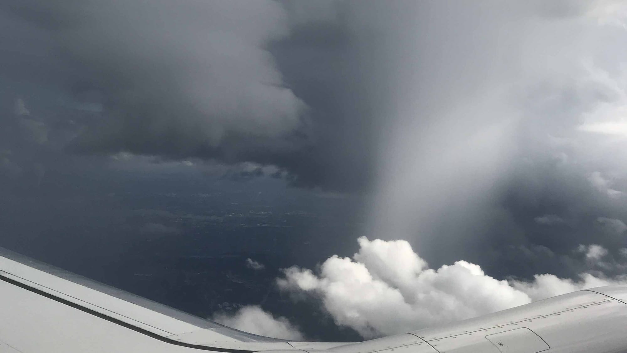 Part of an airplane wing stretches in a shallow U across the bottom of the frame, with puffy white clouds in the U and a blurry gray veil of cloud reaching up from the U to dark gray clouds above, with shadowy terrain showing in the gap between. 