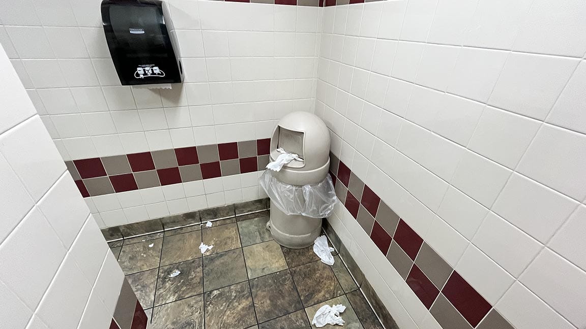 A wide shot of the grimy trashcan in the lavatory, and you can see the paper towel dispenser and more paper towels littering the floor