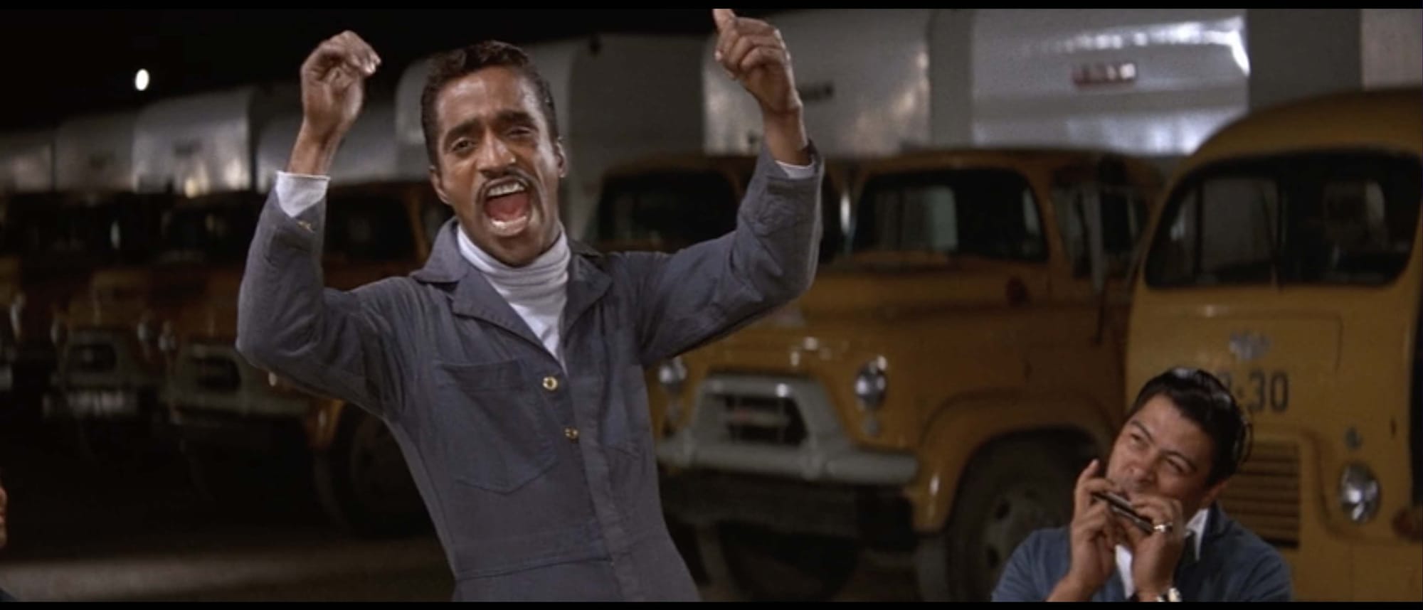 Sammy Davis Jr, shown from the waist up in slim gray coveralls over a white turtleneck, throws up his arms and sings with his mouth wide open. To the right of him are the head, shoulders, and hands of a man in the same outfit playing a tilted harmonica. Behind them, in the dark, a line of garbage trucks with shiny yellow cabs and white bodies recedes from right to left.