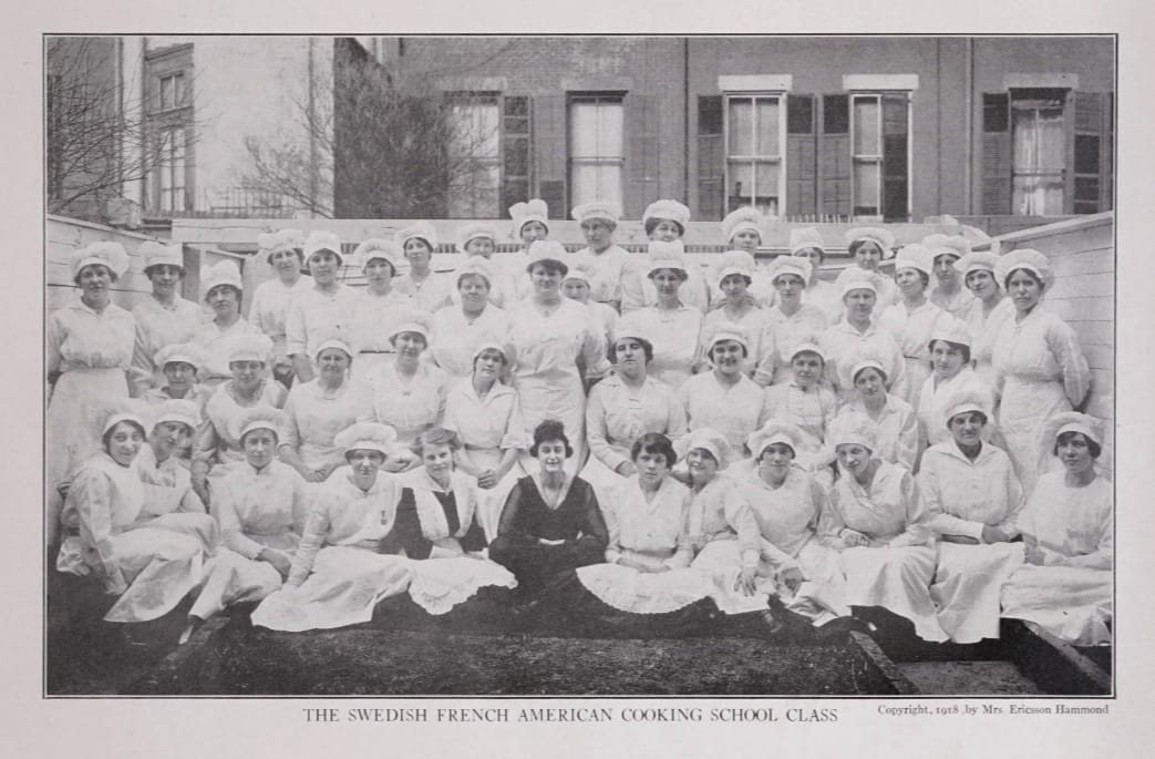 We assume it's Mrs. Ericsson Hammond in dark clothing, center, with an aide to her left in black with a white apron on top, flanked by about 40 students in white aprons and hats