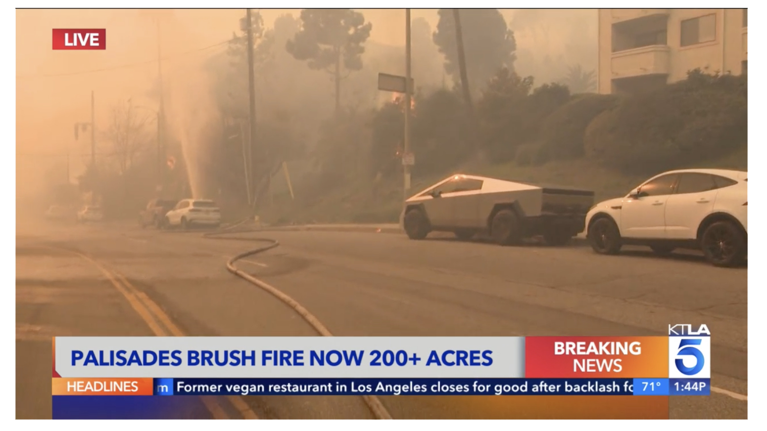 Smoke haze view of a California street with firehose running down street. A geyser of water visible from firehose fitting. Cars parked along street, possibly abandoned, include a Cybertruck