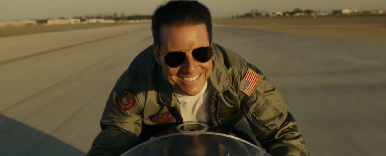 Tom Cruise smiling and wearing aviator sunglasses while riding a motorcycle