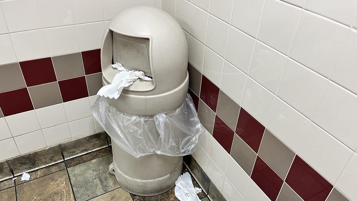 Grimy trashcan in the lavatory with dirty paper towels stuck in the Flap and paper towels on the floor