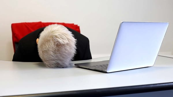 Person at a desk in front laptop computer they are resting their head on desk all you see is top of their head